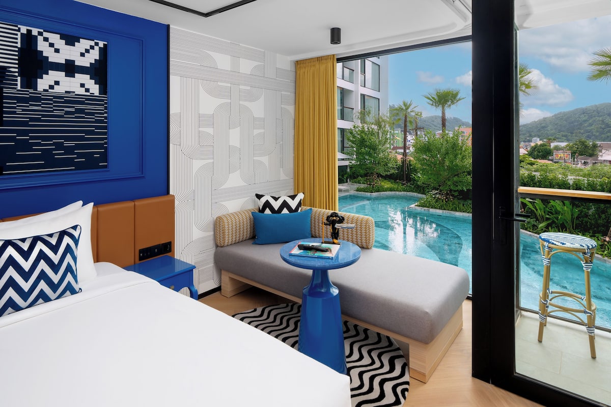 A modern hotel room with a large bed, a sofa, a small table, and a view of a swimming pool and greenery.