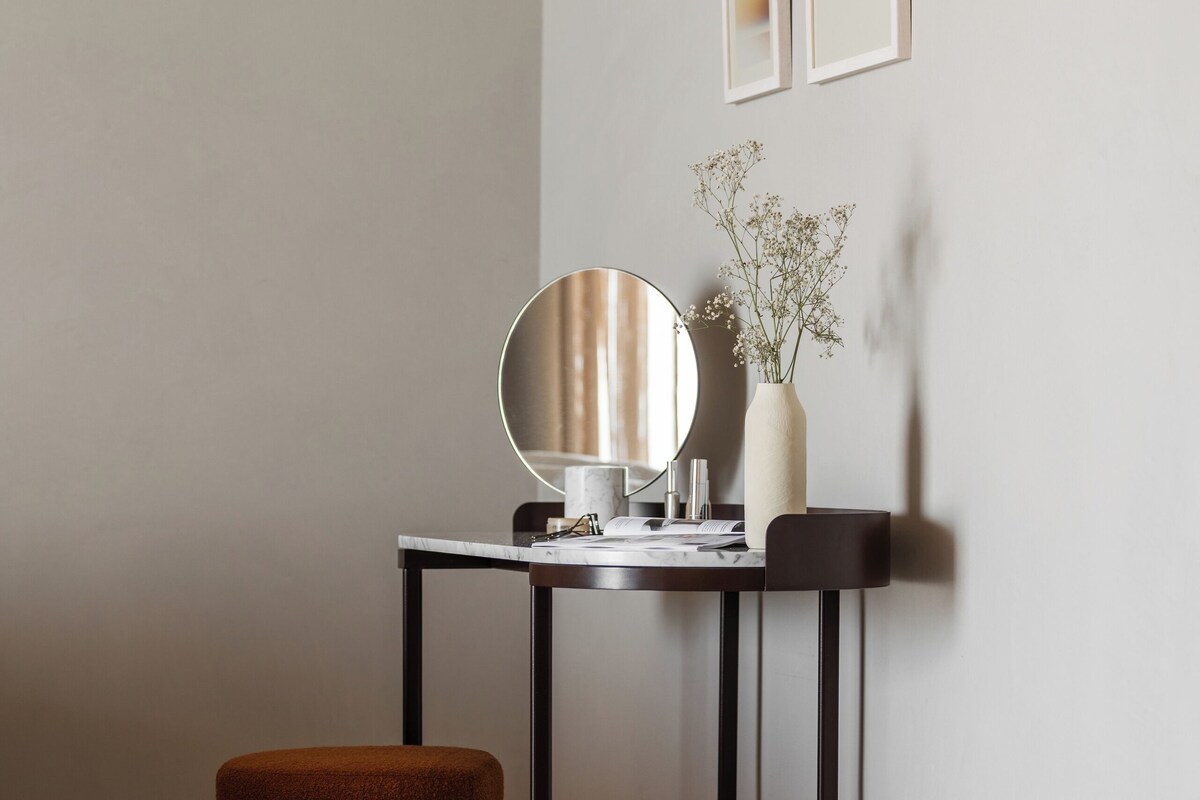 A minimalist room with a round mirror, a small table, a vase with flowers, and a brown ottoman.