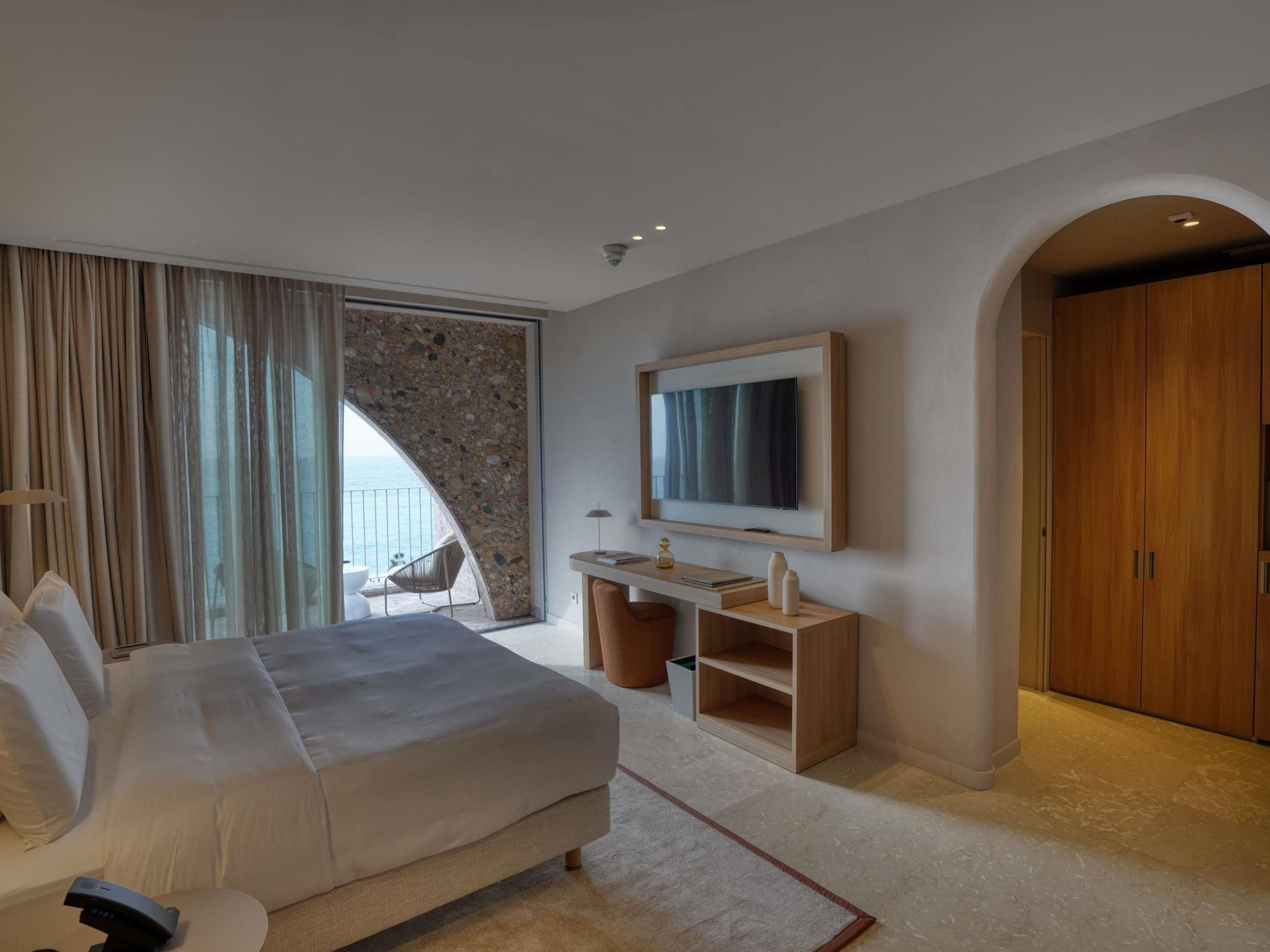 A modern hotel room with a bed, a TV, a desk, and a view of the sea.