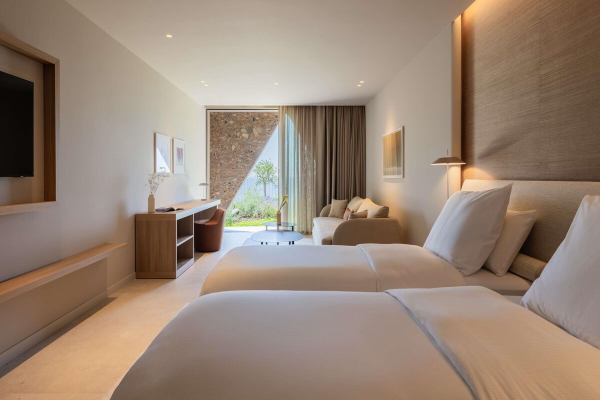 A hotel room with two beds, a desk, a chair, a TV, and a large window with curtains.