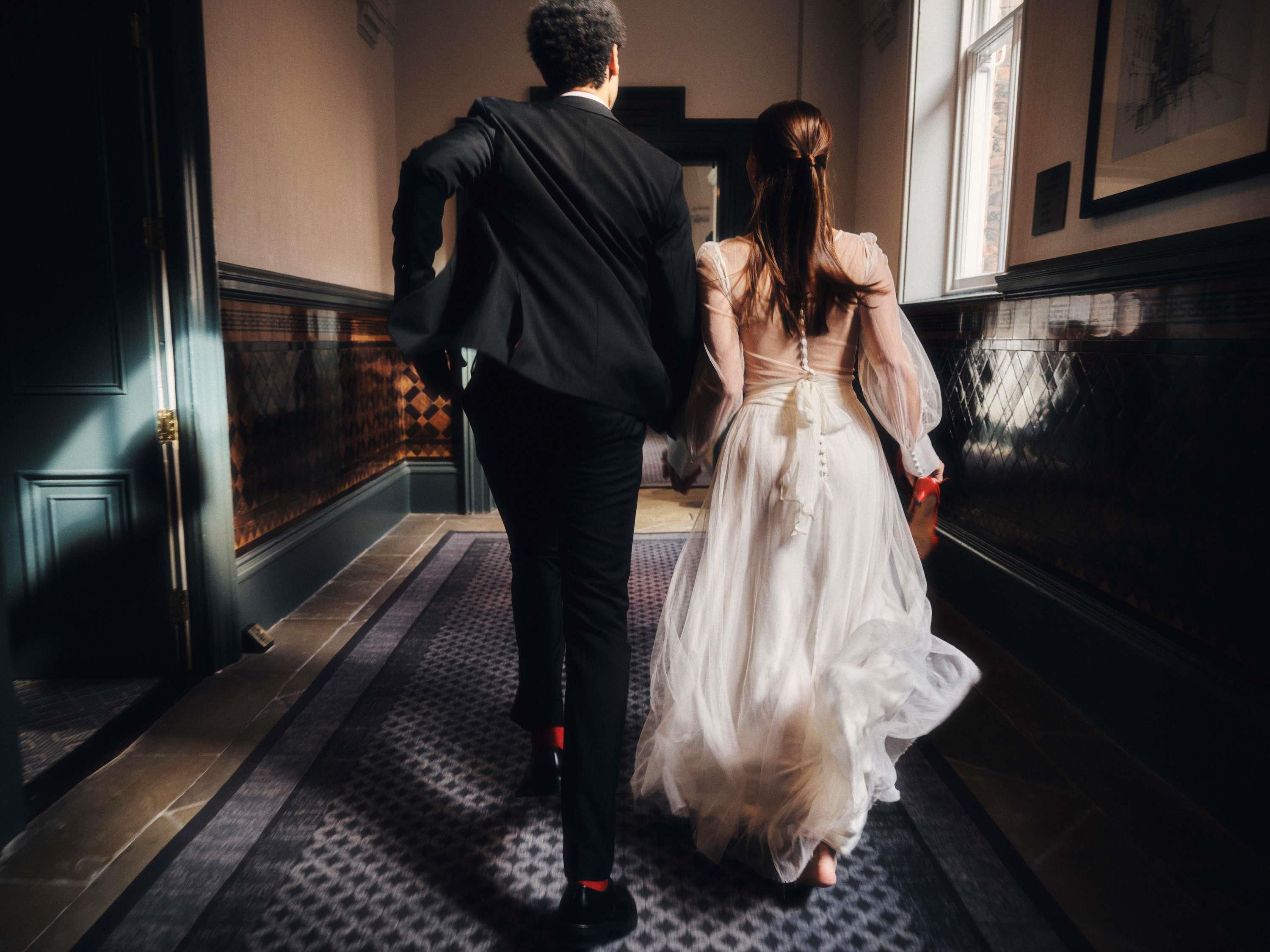 Una pareja camina por un pasillo con alfombra estampada, una puerta a la izquierda y un ventanal a la derecha.