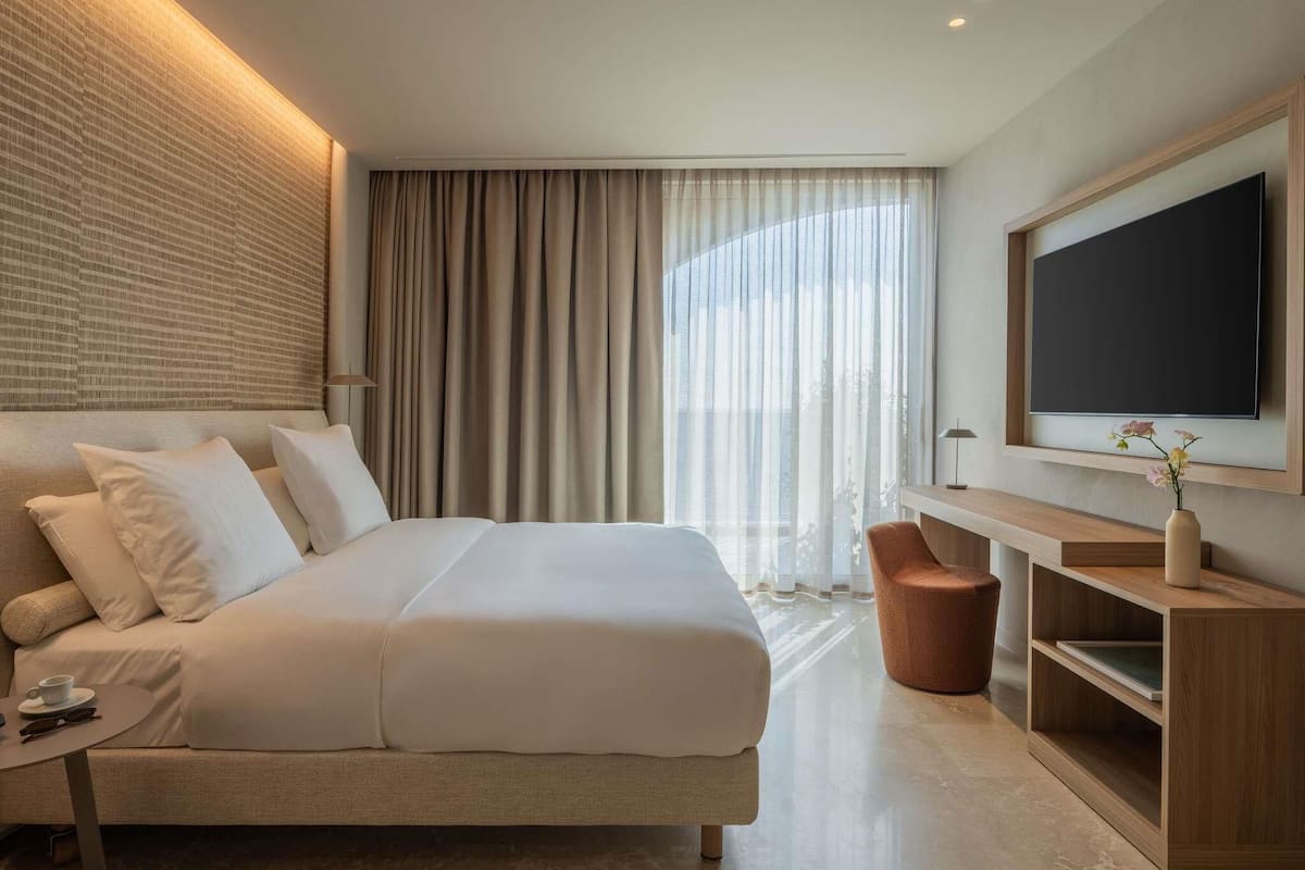 A modern hotel room with a large bed, a TV, a desk, and a view of the outdoors.