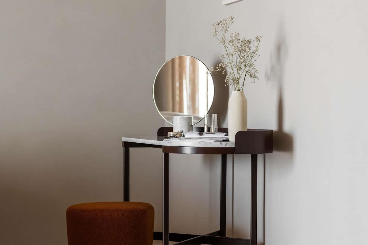 A small, modern vanity with a round mirror, a vase with flowers, and a stool.