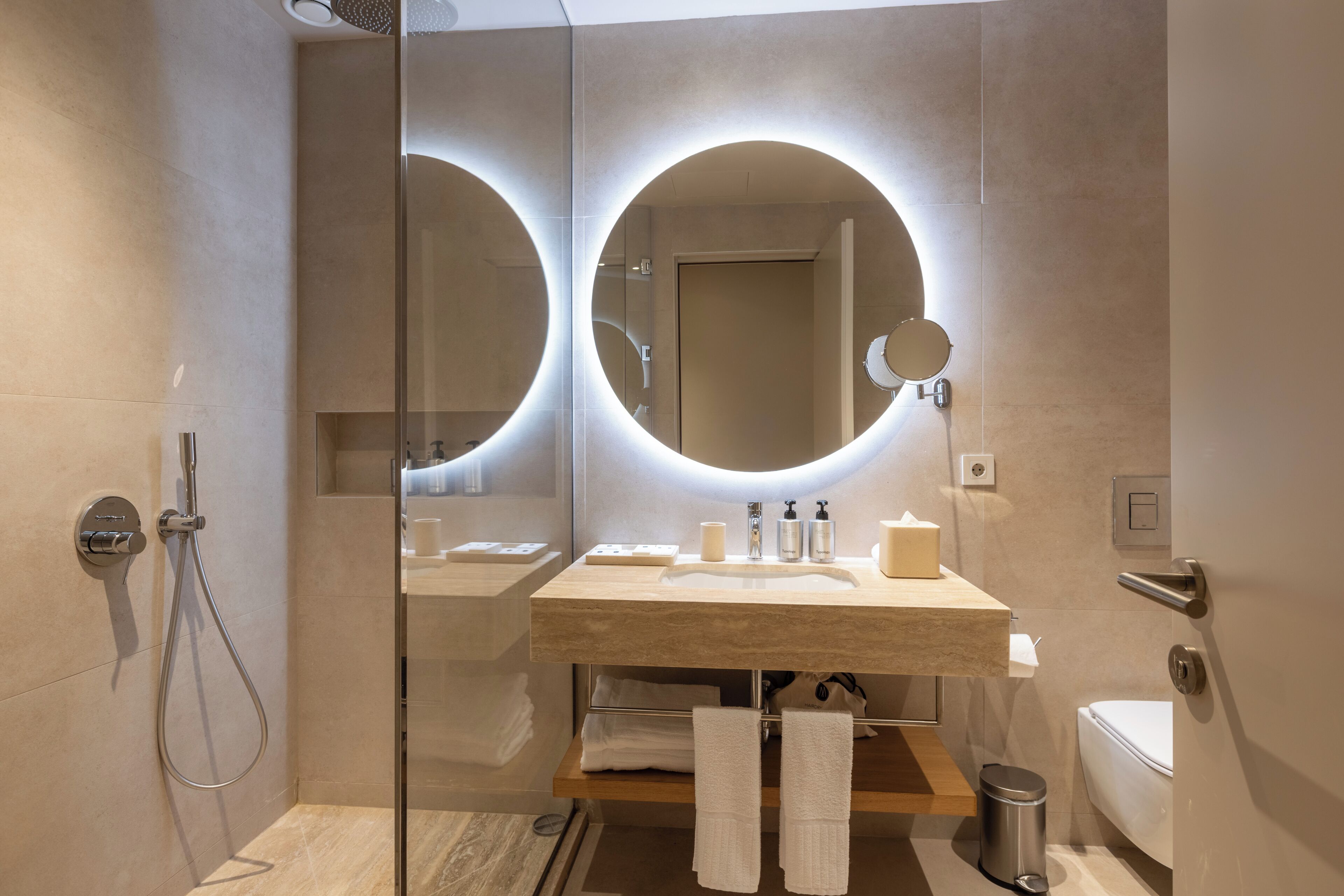 A modern bathroom with a glass shower enclosure, a round mirror, a sink with toiletries, and a toilet.