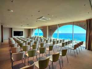 A conference room with rows of chairs facing a large screen, large windows with ocean views, and a flat-screen TV mounted on the wall. - Green Coast Hotel Mgallery Collection (Himare)
