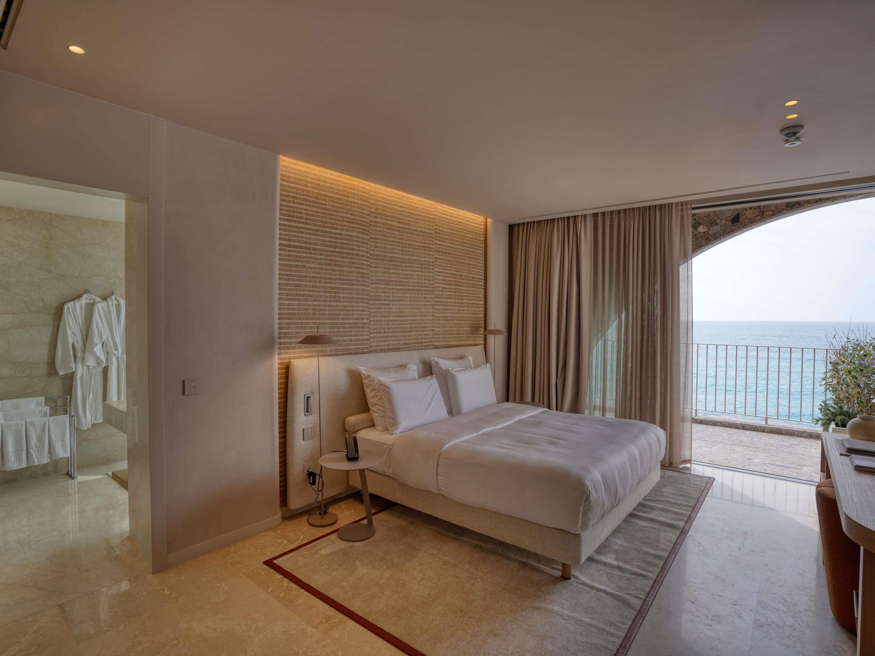 Una habitación de hotel moderna con una cama grande, una mesita de noche, una lámpara y vista al mar.