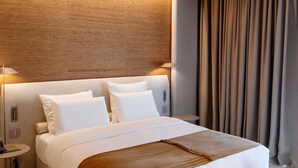 A hotel room with a bed, two bedside tables, a lamp, a curtain, and a wall with a textured pattern. - Green Coast Hotel Mgallery Collection (Himare)