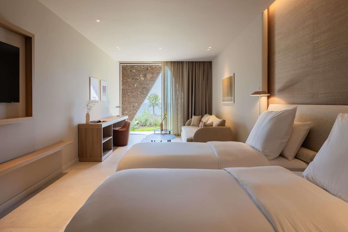 Ein Hotelzimmer mit zwei Betten, einem Schreibtisch, einem Stuhl, einem Fernseher und einem großen Fenster mit Vorhängen.