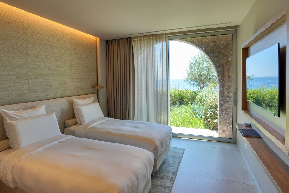 A hotel room with two beds, a large window with a view of the sea, and a flat-screen TV.