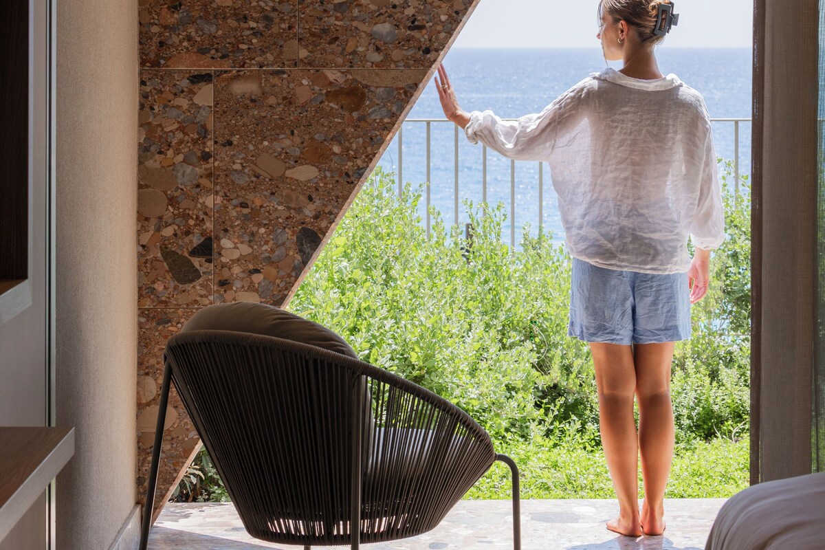 A person standing by a window with a view of the sea, a chair, and a stone wall.