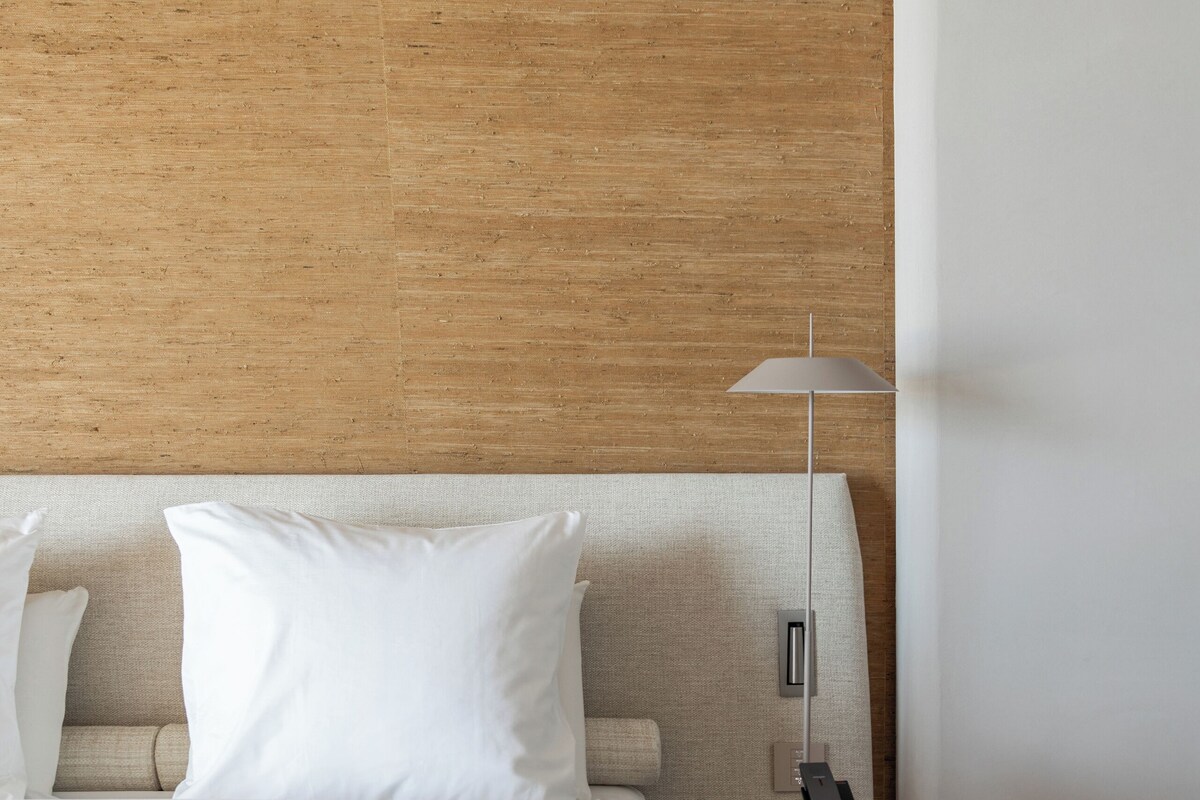A modern hotel room with a wooden headboard, a bed with white linens, a round side table with a lamp, and a small bedside table with a phone.