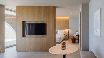 Una habitación de hotel moderna con un televisor de pantalla plana grande montado en un panel de madera, una mesa redonda de mármol con un florero de vidrio, un sofá y una cama con una mesita de noche.