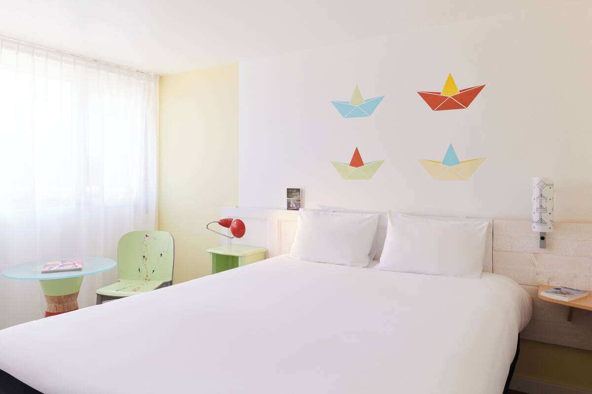 Een hotelkamer met een bed, een tafeltje met een lamp, een stoel en een raam met dunne gordijnen.