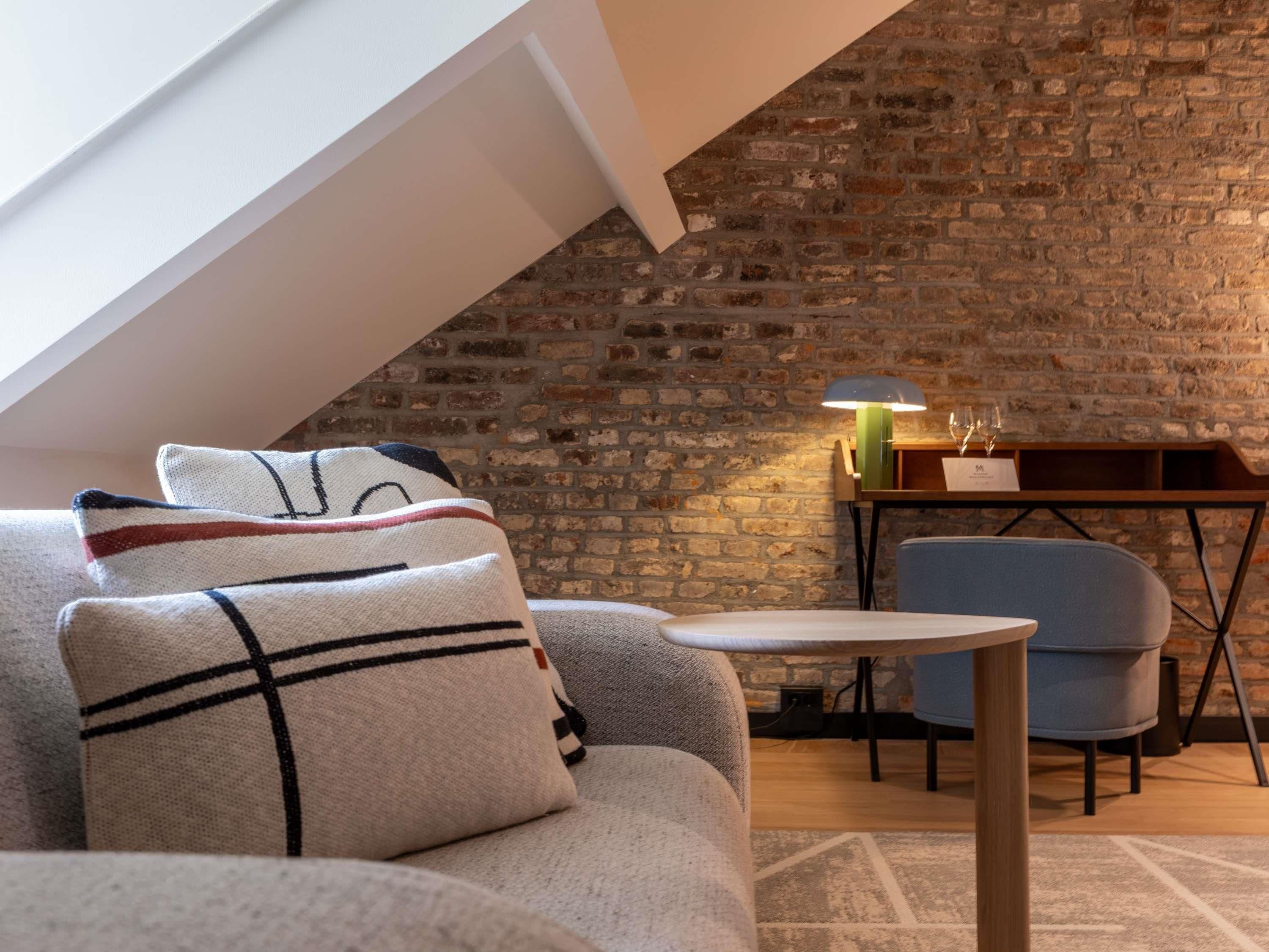 A cozy loft bedroom with a sloped ceiling, exposed brick walls, a modern sofa, a small round table, and a blue chair.