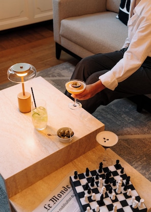 A person holding a cocktail glass, a chessboard, and a menu on a table with a lamp and drinks. - Hotel Maison Maastricht Centrum - Handwritten Collection (Maastricht)