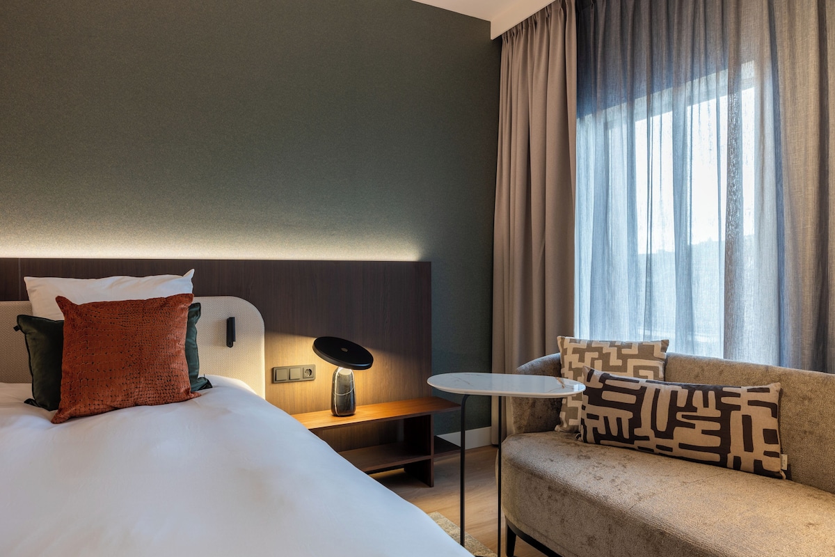 Une chambre d’hôtel avec un lit, une petite table, un canapé et une fenêtre avec des rideaux.