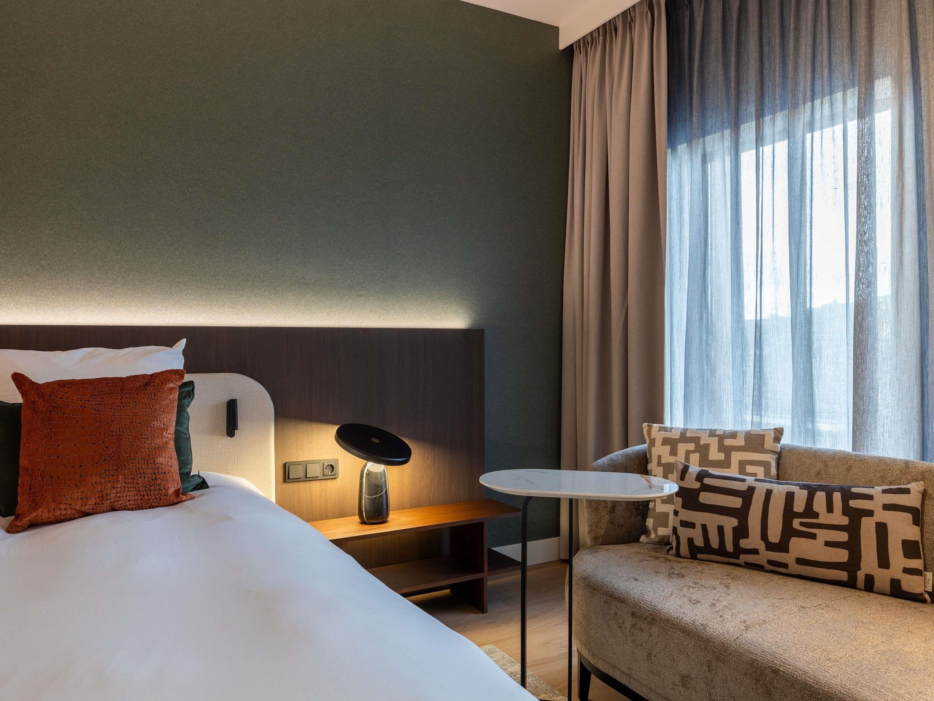 A hotel room with a bed, a bedside table with a lamp, a small round table, and a sofa with patterned cushions.