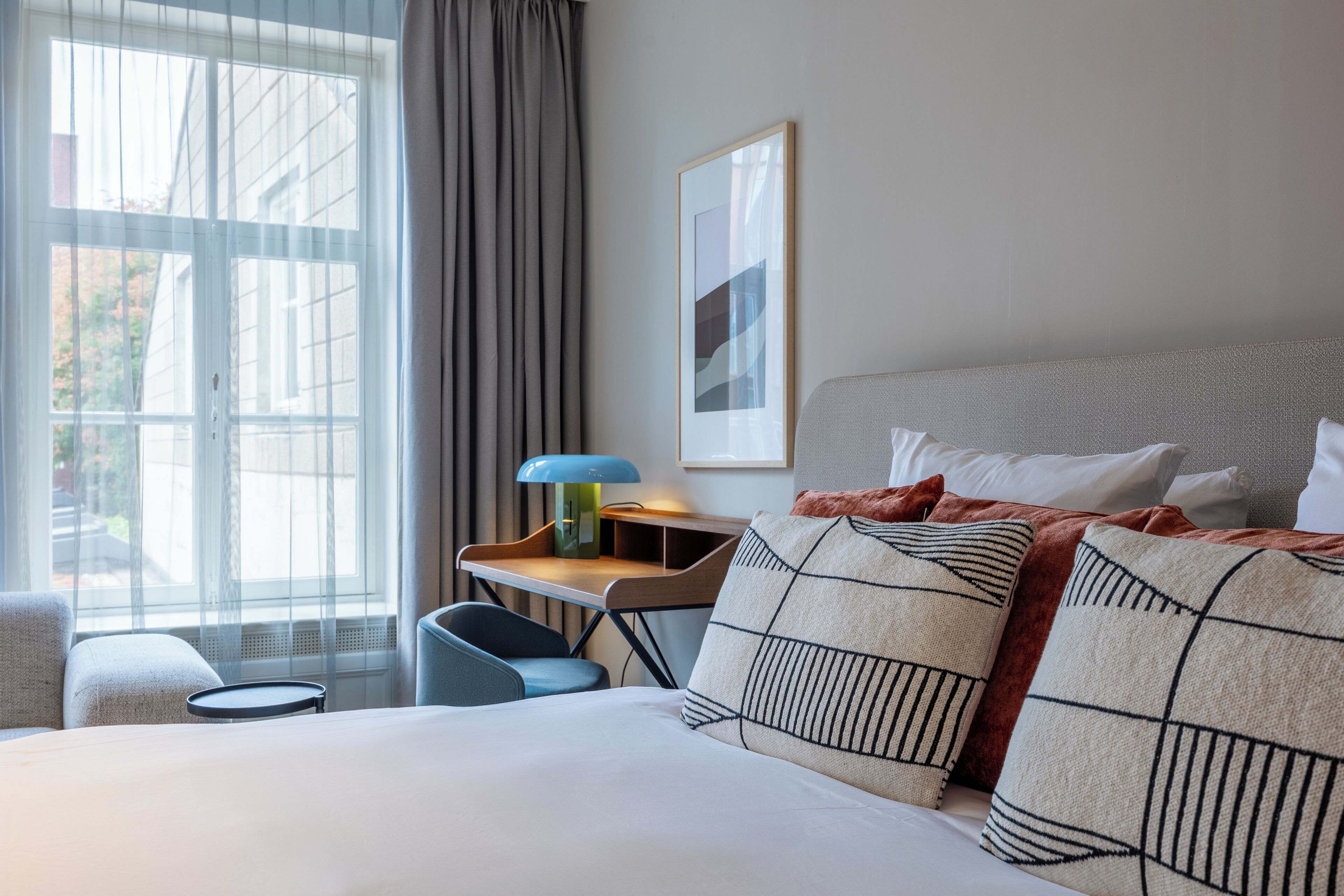 Habitación de hotel con cama, escritorio, silla, ventana con cortinas e imagen en el marco en la pared.