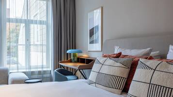 Habitación de hotel con cama, escritorio, silla, ventana con cortinas e imagen en el marco en la pared.