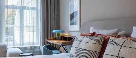 Habitación de hotel con cama, escritorio, silla, ventana con cortinas e imagen en el marco en la pared.