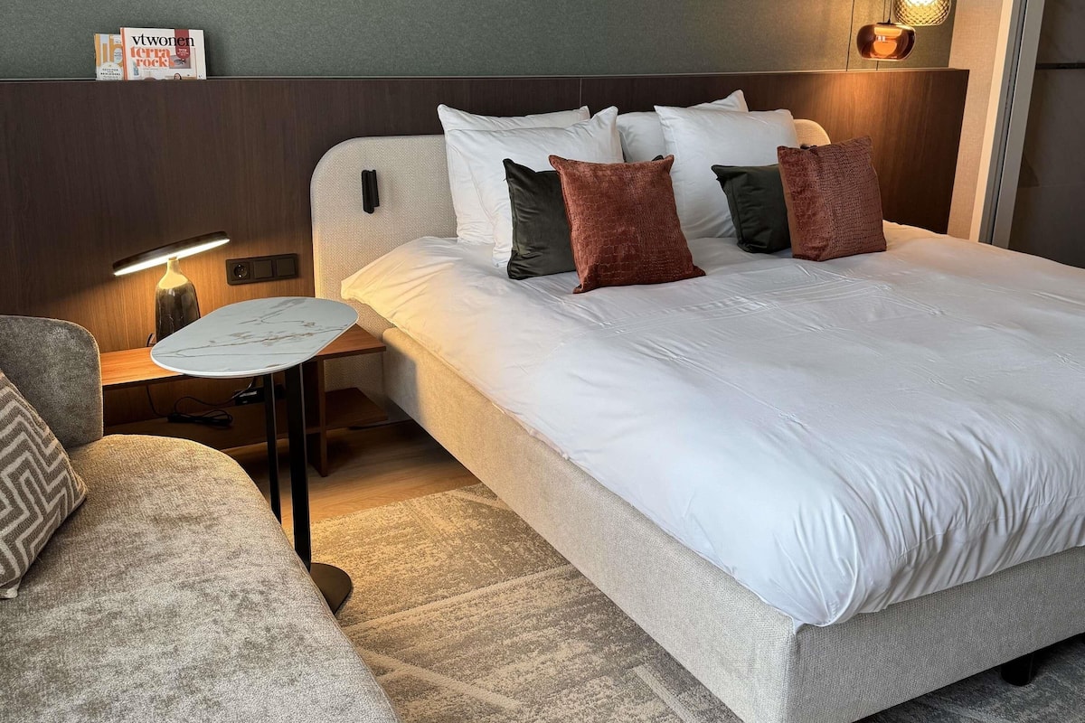 Een hotelkamer met een bed, een kleintje tafeltje, een lamp, een bank en een wandplank met boeken.