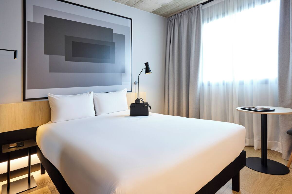 Moderni hotellihuone, jossa on suuri sänky, yöpöytä lampun kanssa, pieni pöytä, jossa on kirja, ja suuri abstrakti maalaus seinällä.