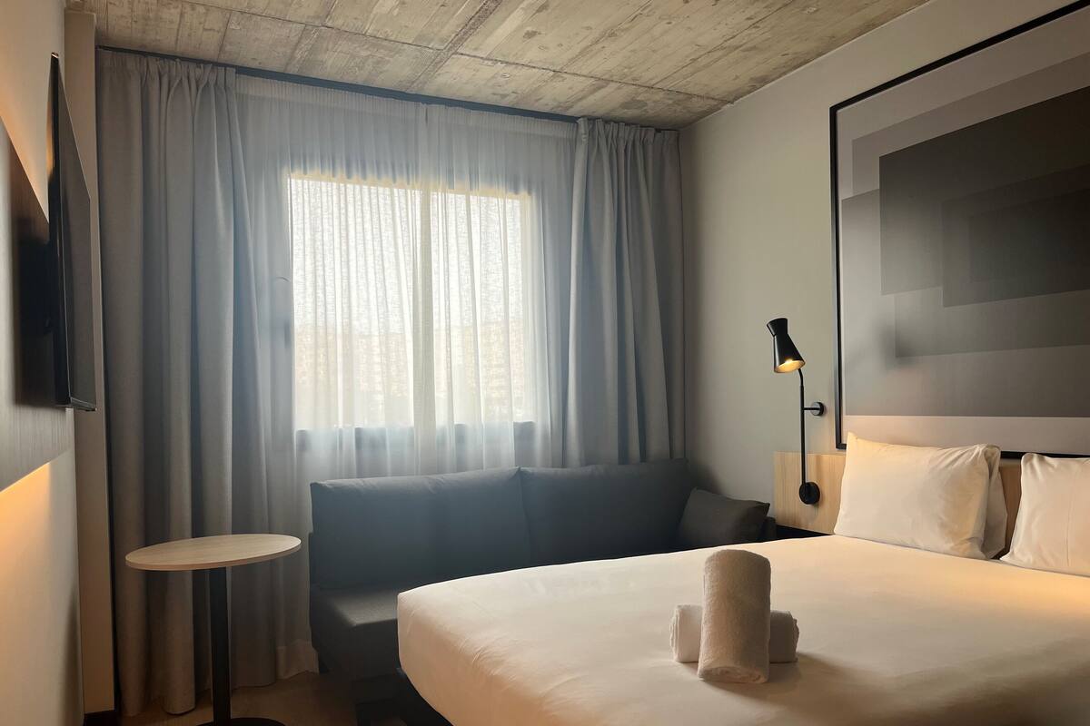 Hotellihuone, jossa on sänky, sohva, pieni pöytä, televisio ja ikkuna, jossa on verhot.