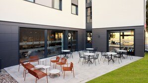 An outdoor patio area with tables and chairs, a grassy lawn, and a building with large windows and a modern design. - ibis Madrid Norte Las Tablas (Madrid)