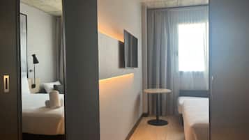 A hotel room with two beds, a small round table, a wall-mounted TV, and a window with curtains.