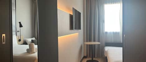 A hotel room with two beds, a small round table, a wall-mounted TV, and a window with curtains.