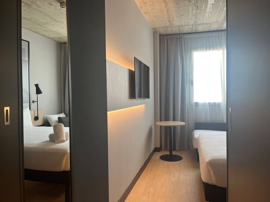 A hotel room with two beds, a small round table, a wall-mounted TV, and a window with curtains.