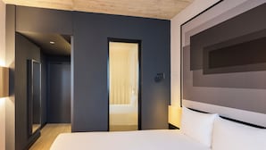 A modern hotel room with a large bed, a door leading to a bathroom, and a wall-mounted light fixture. - ibis Madrid Norte Las Tablas (Madrid)