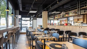 A modern restaurant with long communal tables, individual booths, and a bar area with a variety of bottles and glasses. - ibis Madrid Norte Las Tablas (Madrid)