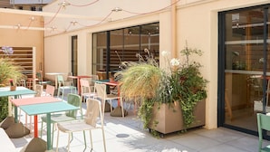 An outdoor seating area with tables and chairs, a large planter with plants, and a building with large windows in the background. - Ibis Styles Marseille Palais Des Congrès Vélodrome (Marseille)