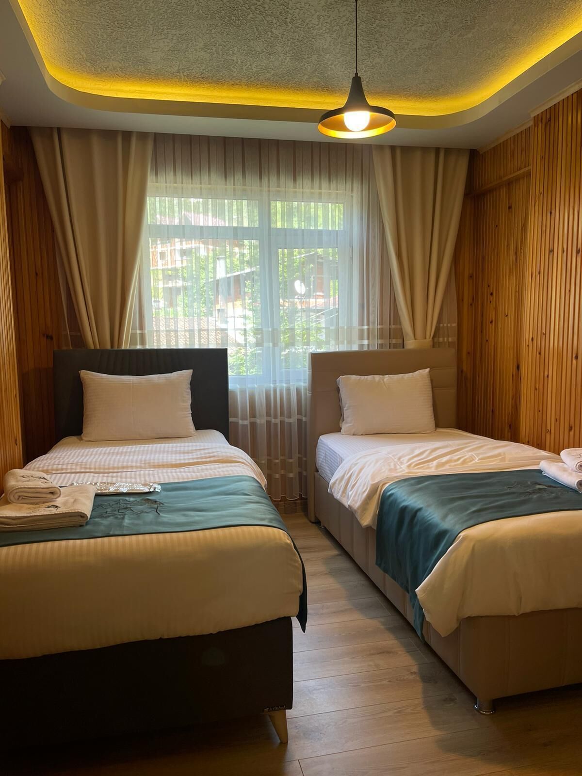 A hotel room with two beds, wooden paneling, a large window with sheer curtains, and a hanging light fixture.