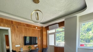 A modern living room with a wooden wall panel, a dining area with a table and chairs, a large window with a view of greenery, and a ceiling light fixture. - EL'A APART HOTEL (Camlihemsin)