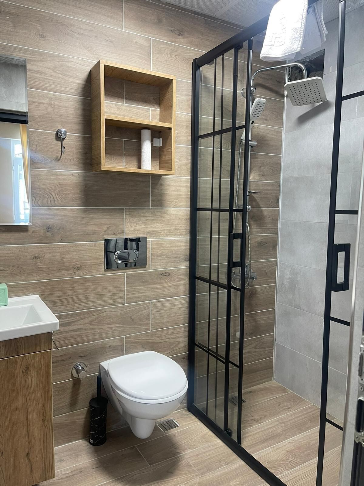 Un baño moderno con suelo de madera, inodoro blanco, cabina de ducha de cristal y estante de madera con una toalla.