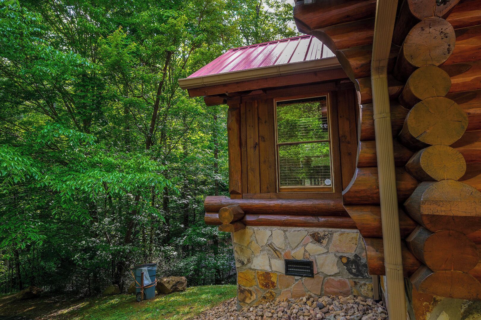A log cabin with a stone foundation, a window with a view of the forest, and a metal roof.
