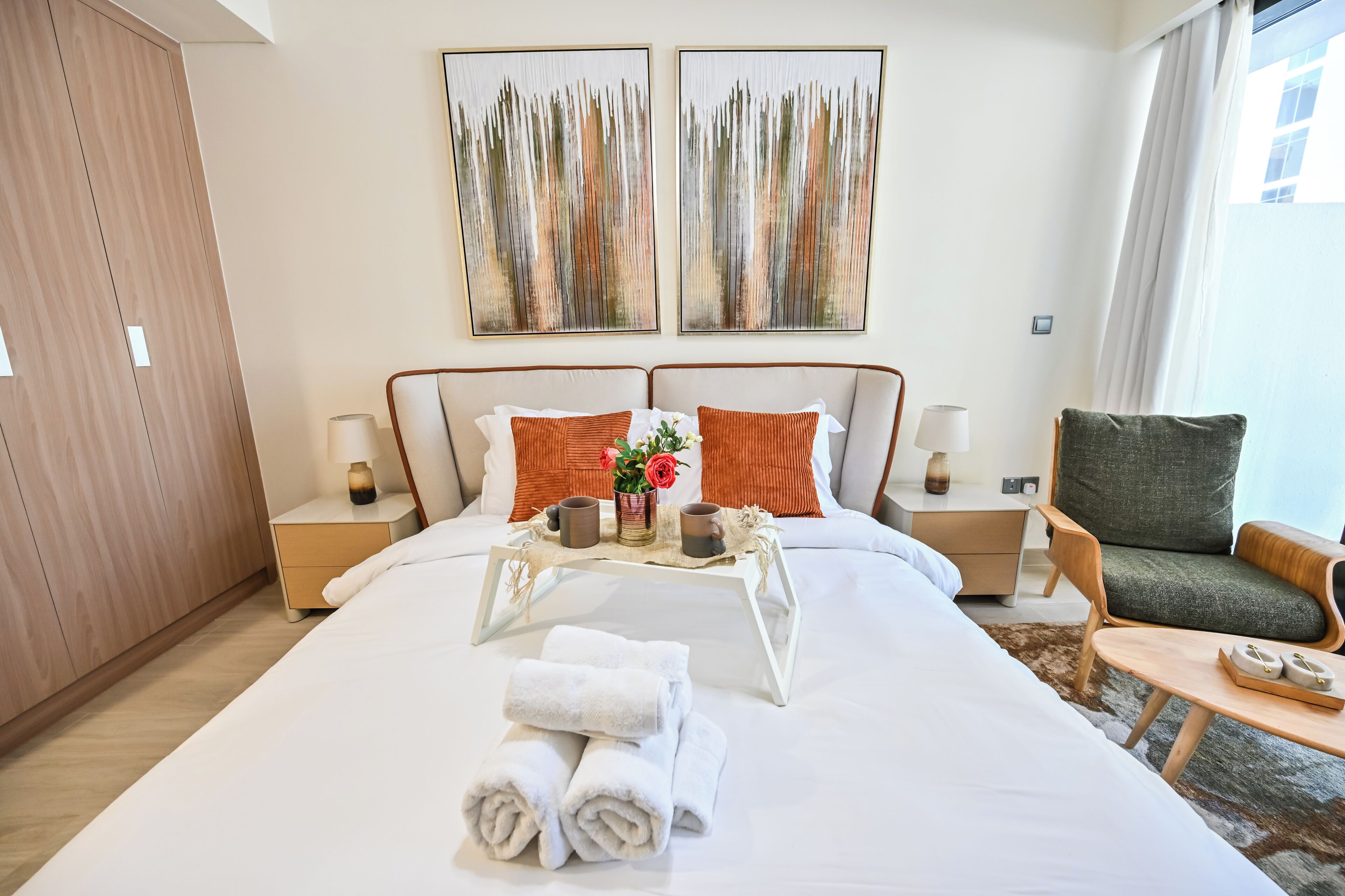 A neatly made bed with towels, a tray with cups and a vase of flowers, and a chair by the window.