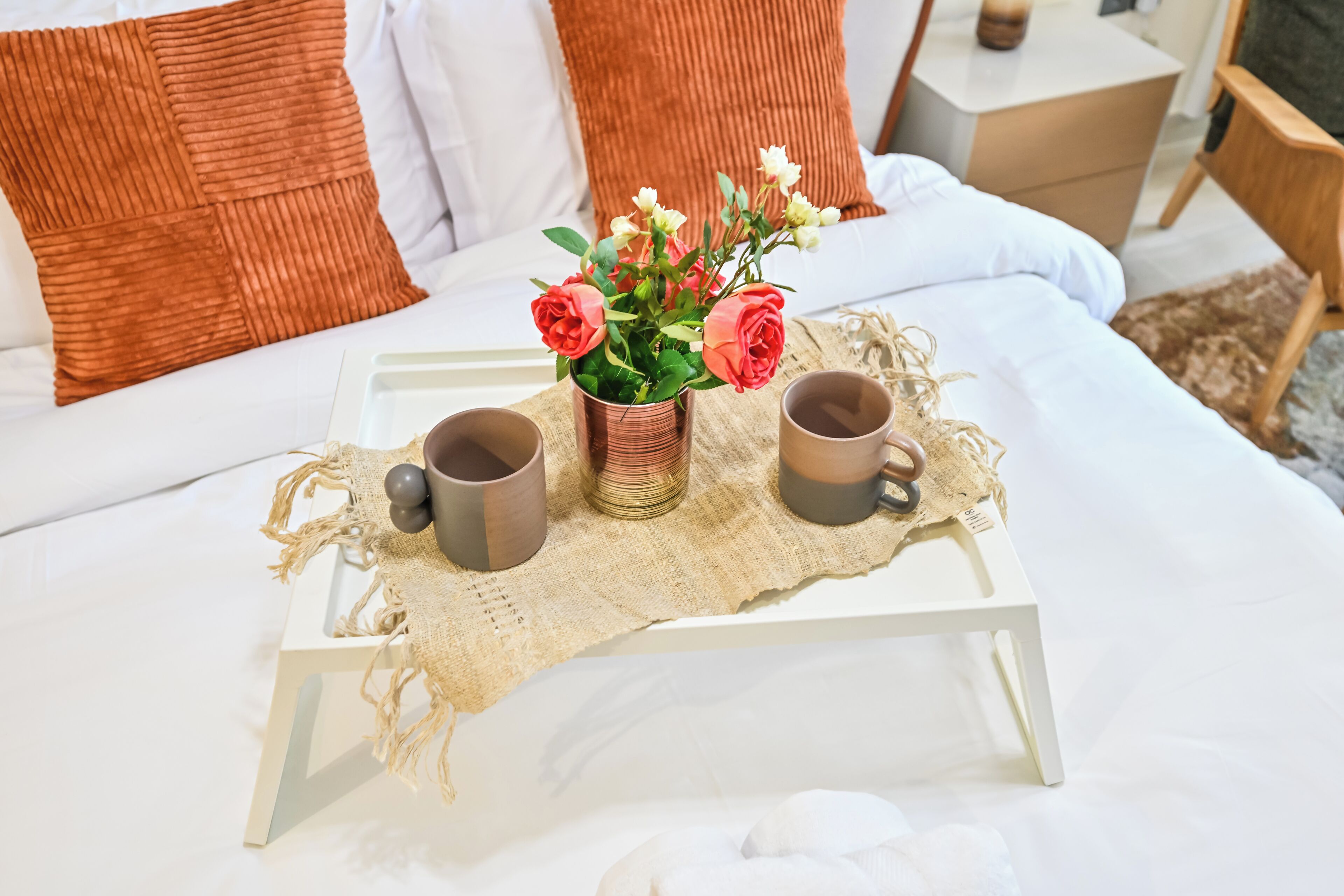 Uma cama bem arrumada em tons de branco e castanho, um pequeno criado-mudo branco com uma luminária, e uma bandeja com duas canecas e um vaso de flores.