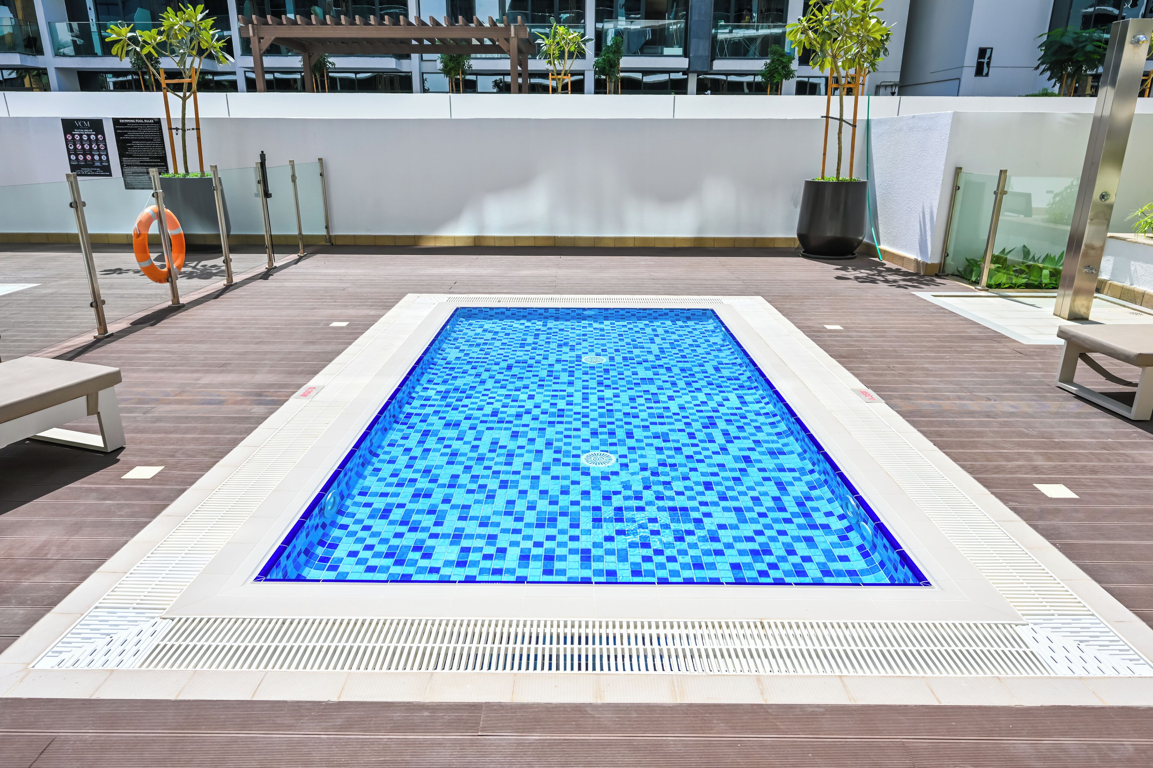 A rectangular pool with a blue mosaic tile pattern, surrounded by a wooden deck and a white wall with a sign.
