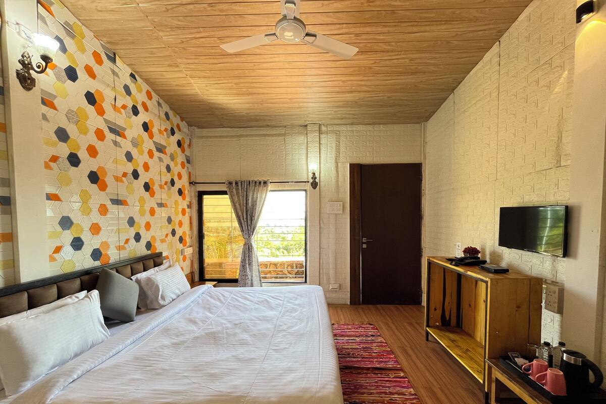 A bedroom with a large bed, a wooden ceiling fan, a TV on a wooden stand, and a view of the outdoors through a door.
