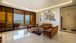A spacious living room with a large glass door leading to a balcony, a sofa, a coffee table, and a rug. - Astoria Lagoon Resort (Igatpuri)