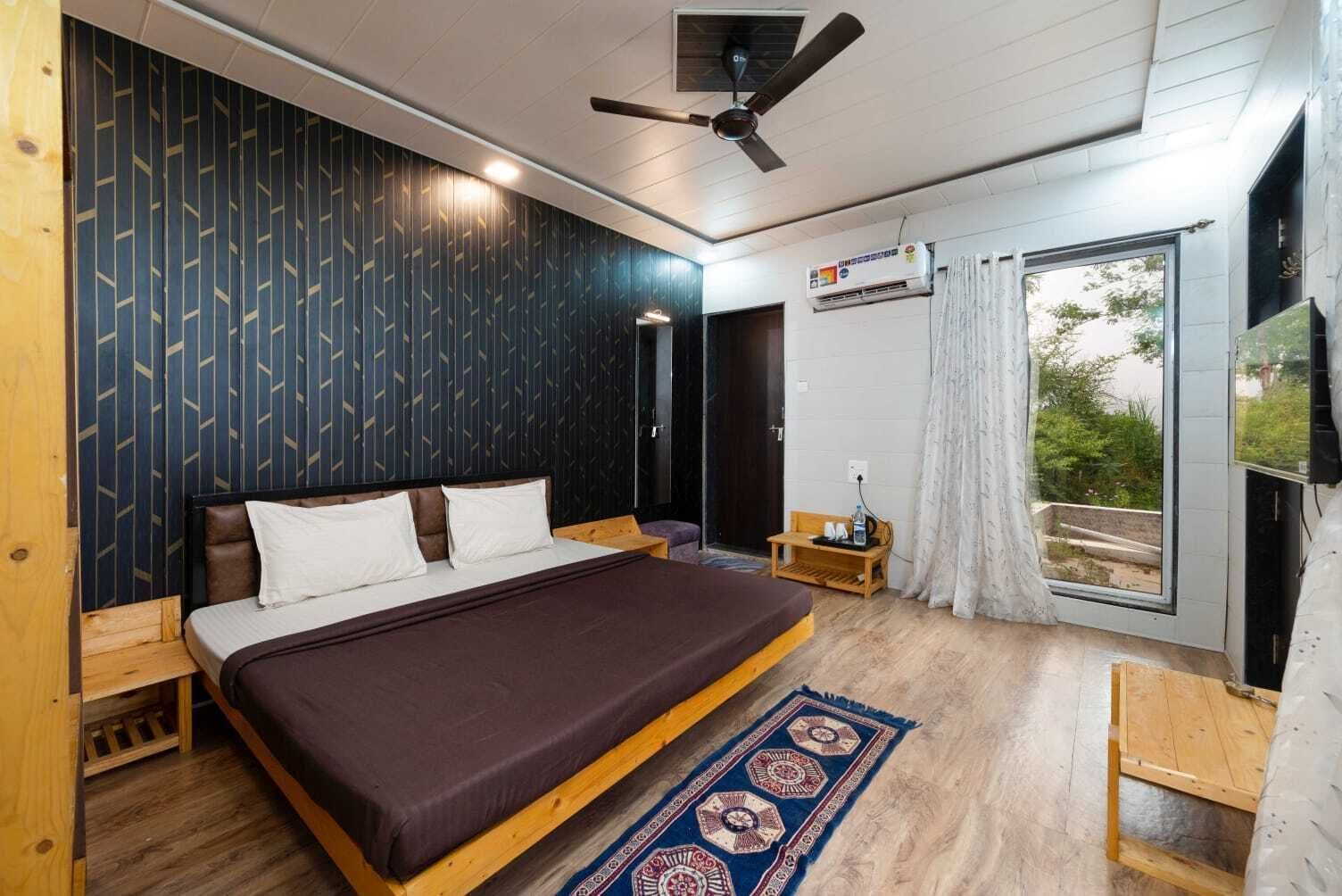 Un dormitorio con una cama grande, un ventilador de techo, una ventana con cortinas y una pequeña mesa con una botella.