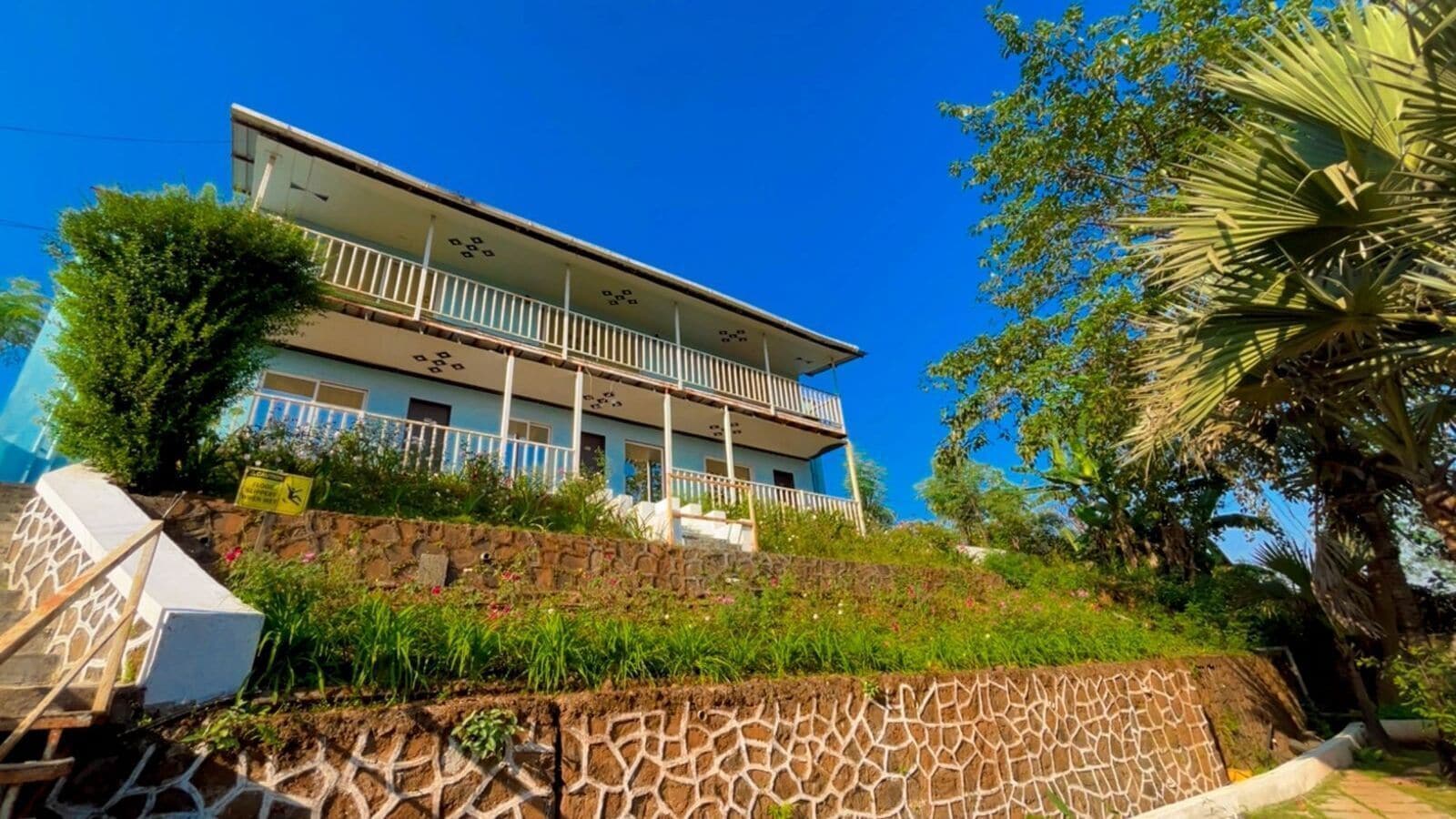 A two-story building with a balcony, surrounded by lush greenery and a stone retaining wall.