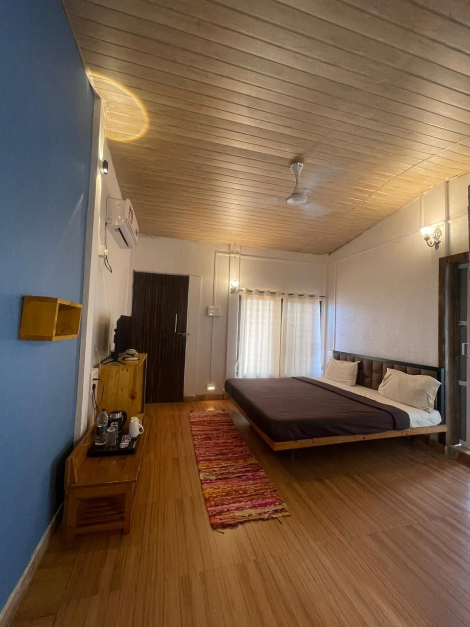 A bedroom with a bed, wooden floor, a small table with items, a ceiling fan, and a window with curtains.