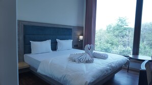 A hotel room with a large bed, a headboard, a nightstand, a lamp, and a view of trees through the window.