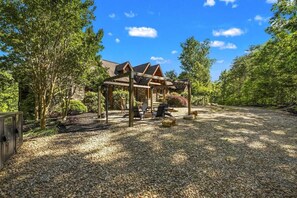 A covered outdoor seating area with a stone-paved ground, surrounded by trees and a building with a sloped roof.