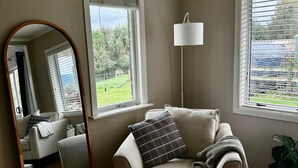 A living room with a beige sofa, a floor lamp, a large mirror, and a view of a garden through the windows. - Ruby’s Nest – A countryside experience (Te Poi)