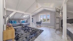 A spacious loft bedroom with a large window, a grey sofa, a rocking chair, and a decorative rug.